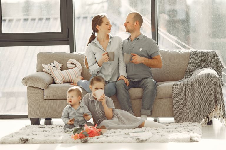 Happy family with children spending time at home while parents sitting on couch with cups of coffee and looking at each other when kids resting on floor