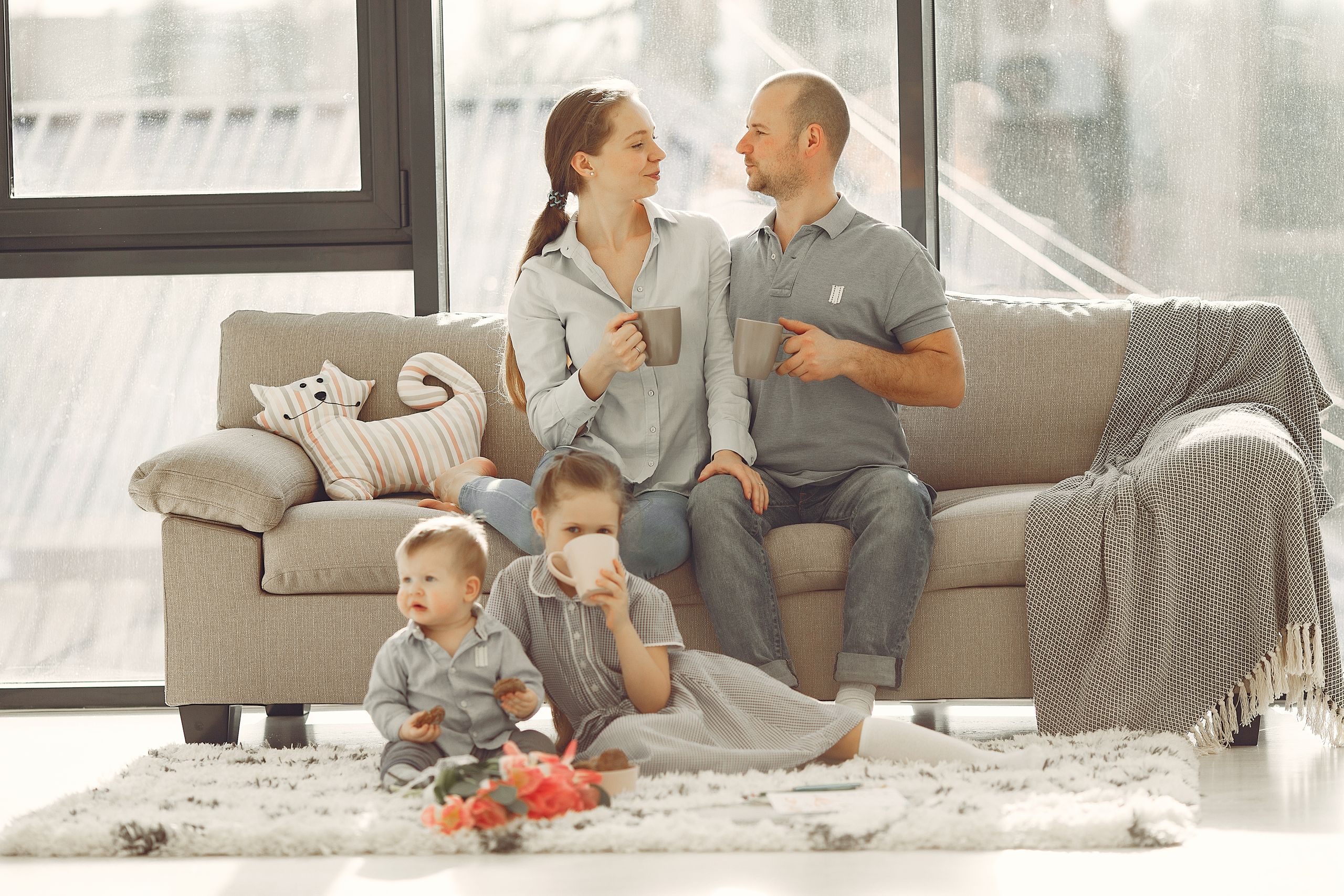 Happy family with children spending time at home while parents sitting on couch with cups of coffee and looking at each other when kids resting on floor
